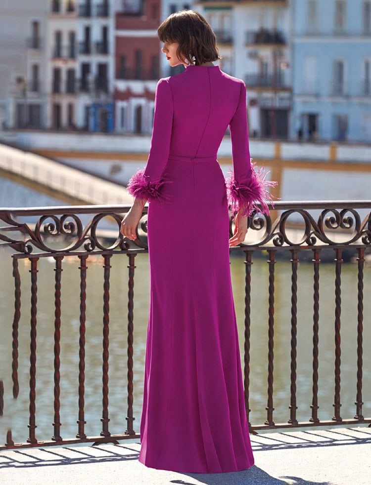 Femme portant une robe longue violette avec des détails en plumes sur les manches, vue de dos sur un pont avec vue sur des bâtiments colorés.