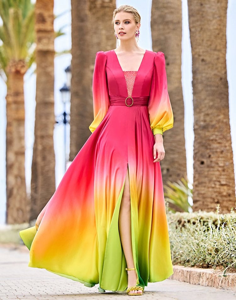 Une femme portant une robe longue fluide dégradée de rose, jaune et vert, avec un décolleté en V et une ceinture dorée, dans un décor en plein air avec palmiers.