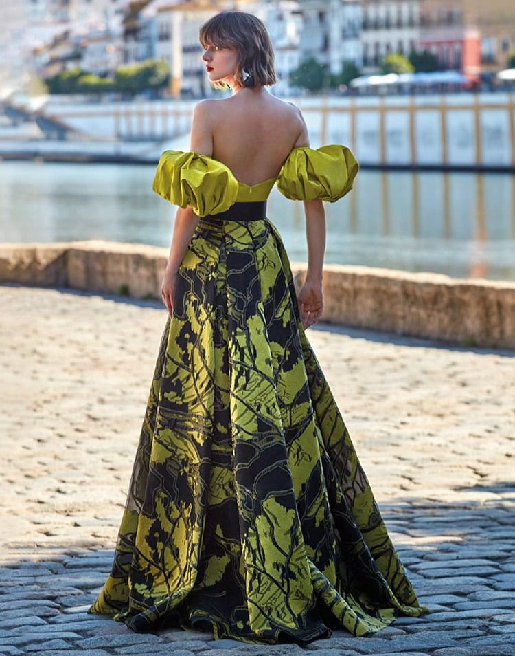 Robe de soirée longue avec épaules dénudées et manches bouffantes, motif floral vert et noir, portée par une femme près d'un port, style chic et sophistiqué.