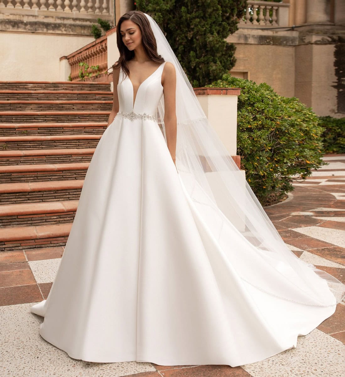 Un voile de mariée blanc élégant avec robe de mariage longue et décolleté en V, portée par une femme dans un lieu historique en plein air, entourée de verdure et de marches en pierre.