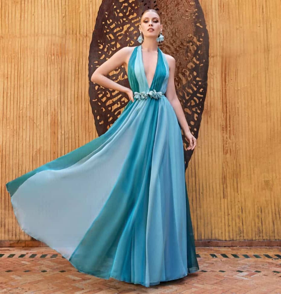 Élégante femme portant une robe longue bleue avec décolleté en V, ceinturée à la taille, posant devant un fond en bois avec un motif en filigrane noir.