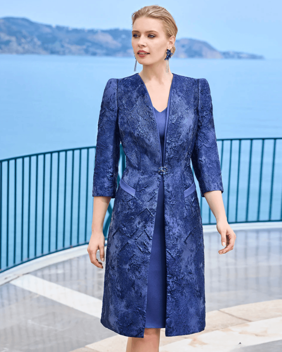Femme portant une robe longue en tissu texturé bleu marine, avec un manteau assorti, en extérieur près de la mer, style sophistiqué et moderne.
