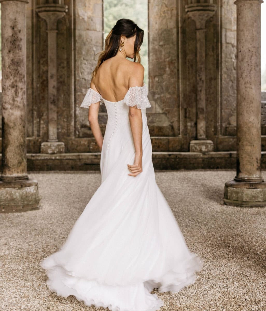 Robe de mariée blanche élégante avec épaules dénudées et détails en dentelle, posant dans un ancien site historique avec colonnes en pierre.