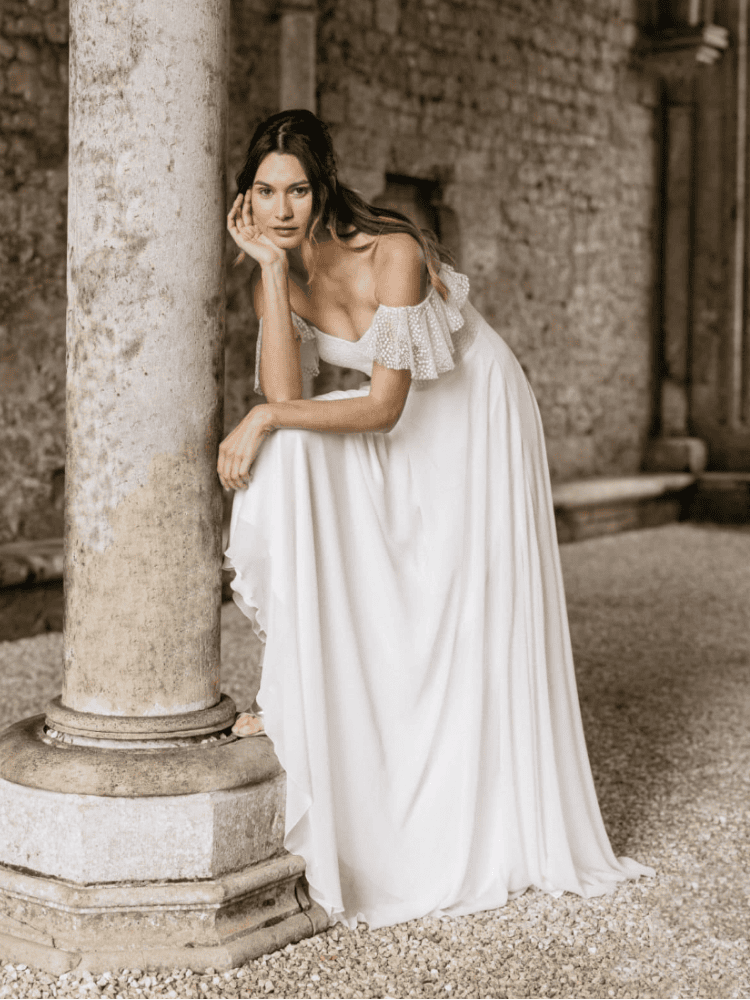 Une femme élégante en robe blanche longue posant près d'une colonne en pierre dans un site historique, mettant en valeur la mode bohème chic et le style sophistiqué.