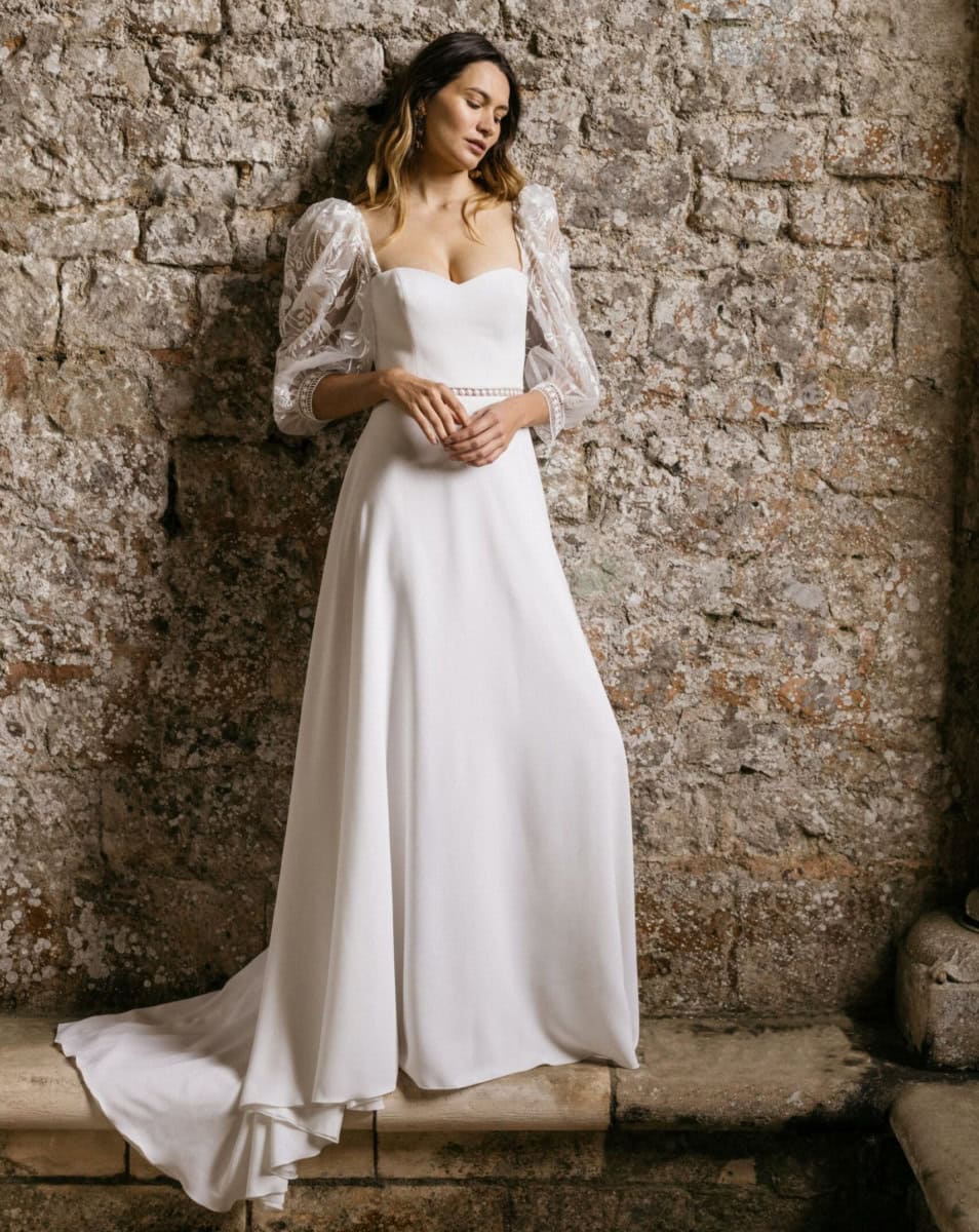 Robe de mariée élégante avec manches en dentelle et corsage en satin, posant contre un mur en pierre pour une séance photo romantique et sophistiquée.