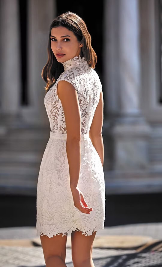 Jeune femme élégante en robe blanche en dentelle, posant devant un bâtiment historique en plein jour.