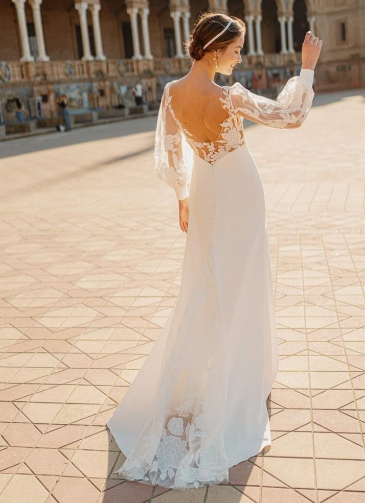 Robe de mariée élégante avec dentelle et manches longues, portée par une femme dans un lieu historique en plein air, ambiance ensoleillée et sophistiquée.