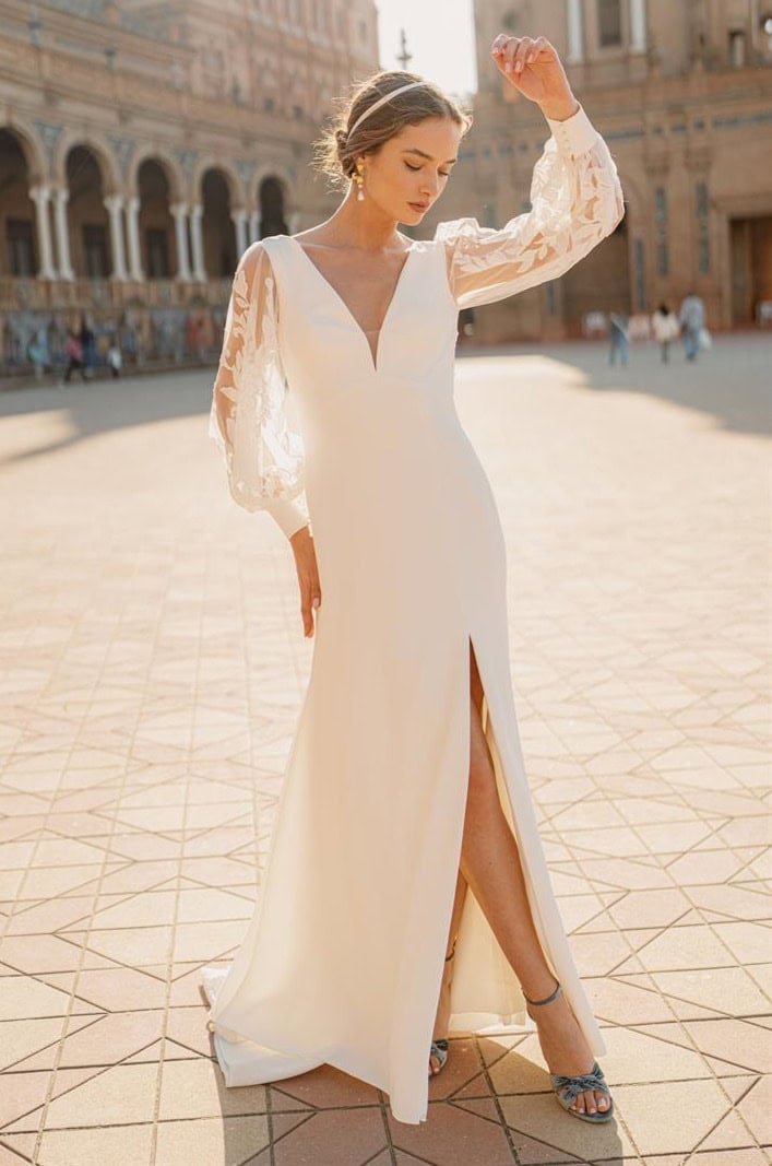 Élégante femme en robe de mariage blanche avec manches en dentelle, posant dans une place historique ensoleillée, mettant en valeur la mode nuptiale et le style sophistiqué.