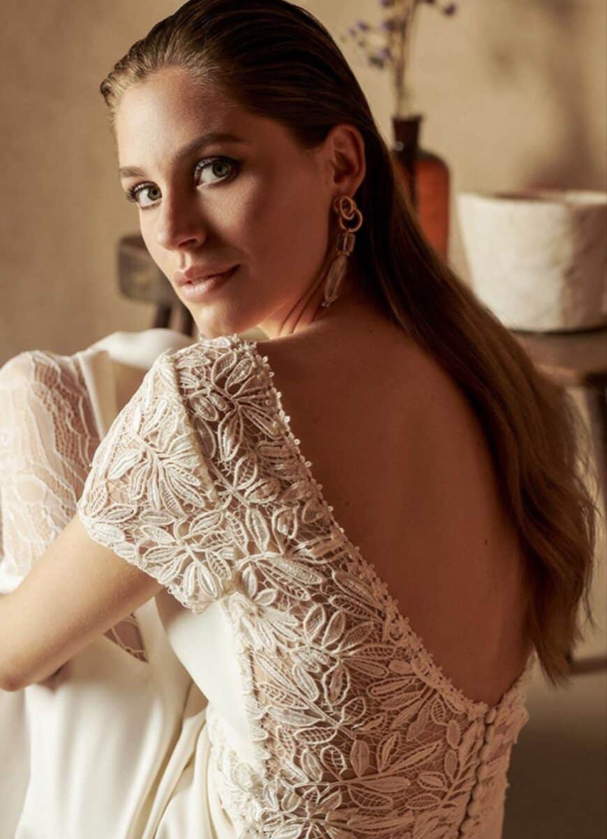 Femme élégante portant une robe en dentelle blanche avec un dos nu, accessoires sophistiqués, dans un intérieur chaleureux et chic, mettant en valeur la mode féminine et le style glamour.