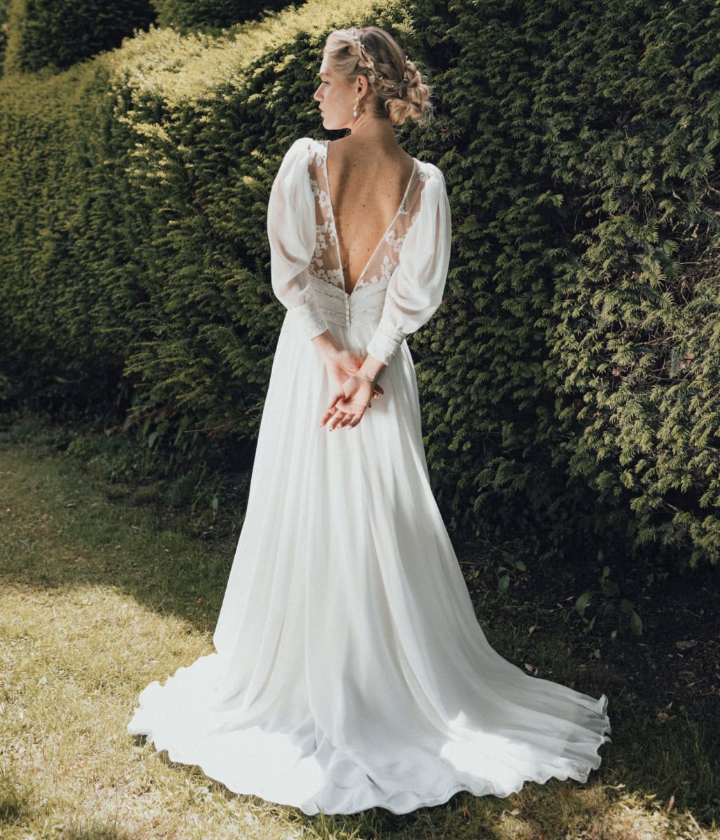 Tage de mariage en robe blanche élégante avec manches bouffantes et décolleté en V, posant dans un jardin verdoyant, mettant en valeur un style romantique et sophistiqué.