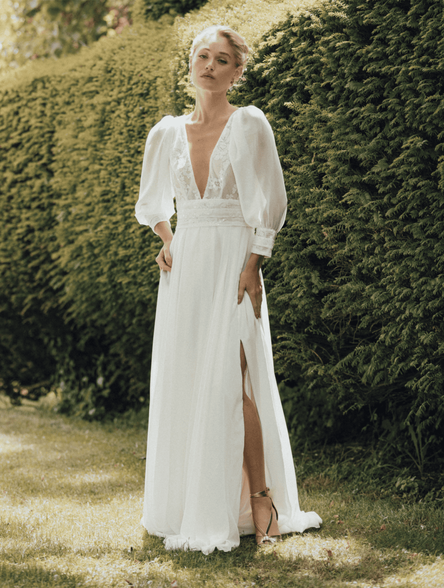 Robe de mariée élégante en tissu blanc avec décolleté en V, manches bouffantes et fente haute, portée par une femme dans un jardin verdoyant, style bohème chic, parfait pour mariage ou occasion spécia.