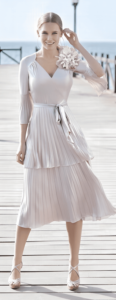 Femme souriante portant une robe d'été en mousseline blanche avec ceinture à nouer, debout sur une jetée en bois avec vue sur la mer en arrière-plan.