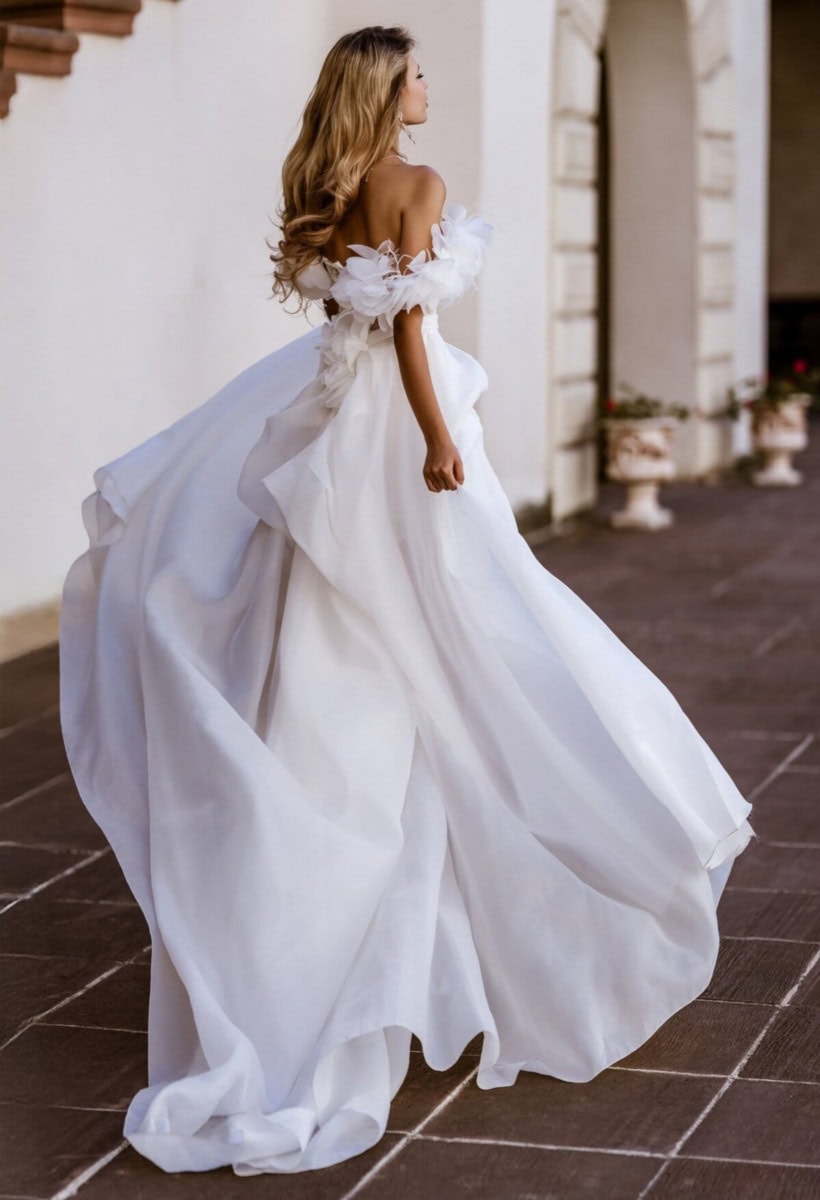 Femme portant une robe de mariée blanche élégante avec détails en tulle et volants, marchant dans une cour en pierre, vue de profil.