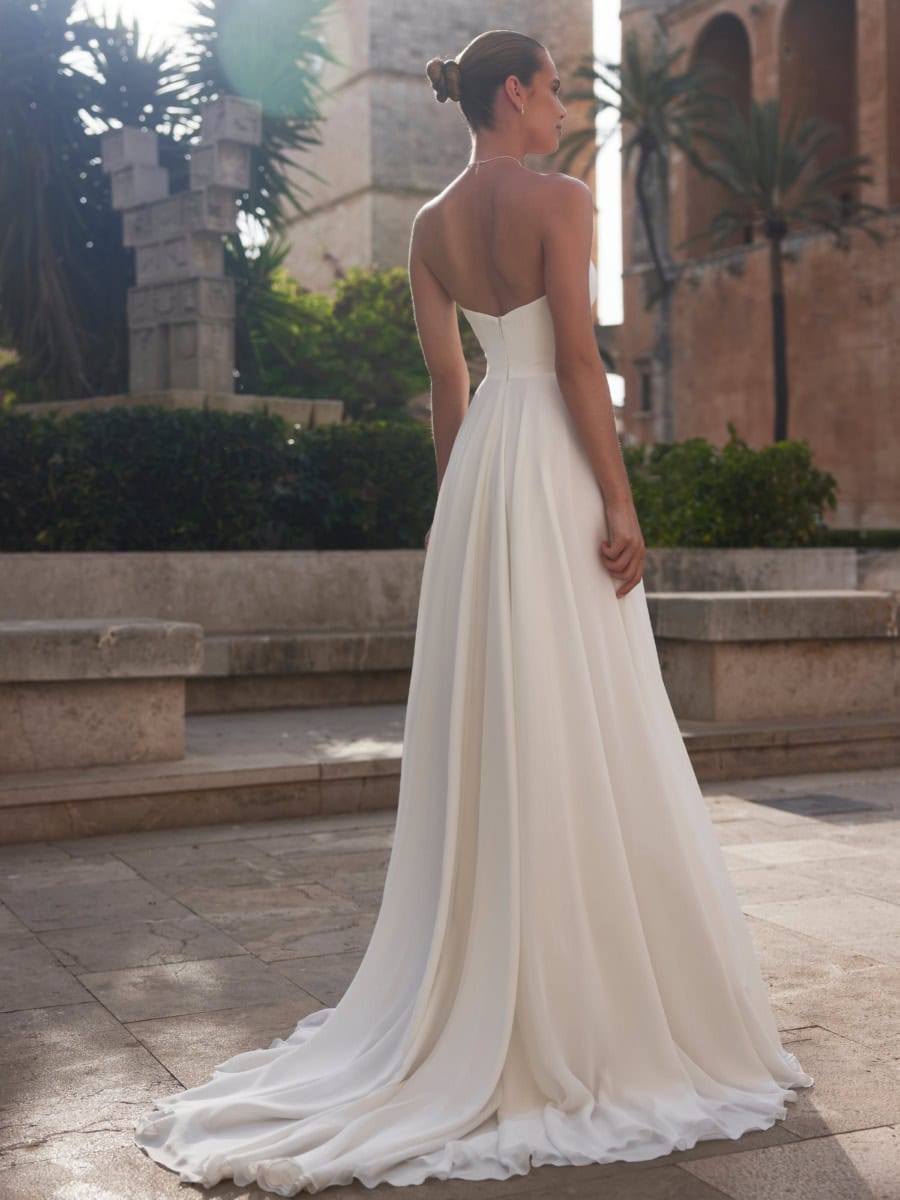Élégante robe de mariée blanche en satin avec dos nu et longue traîne, portée par une femme dans un lieu historique en plein air, entourée de végétation luxuriante et d'architecture ancienne.