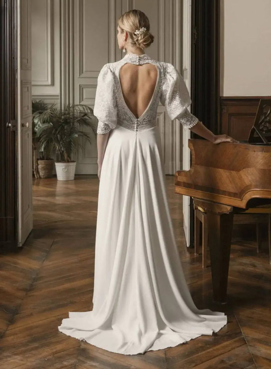 Robe de mariée élégante avec dos nu en satin blanc, manches bouffantes en dentelle, dans un intérieur classique avec piano et parquet en bois.