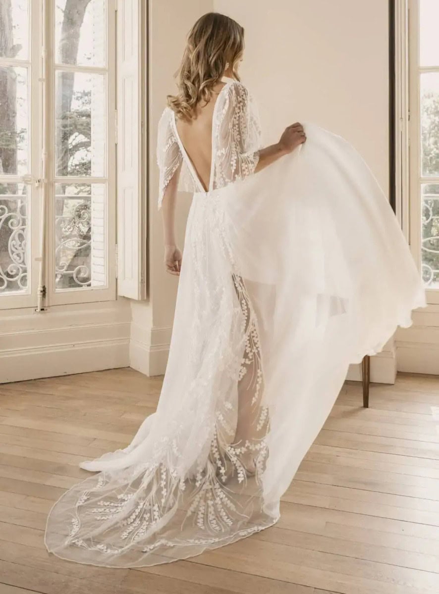 Robe de mariée élégante en dentelle blanche avec dos en V et traîne longue, dans une pièce lumineuse avec grandes fenêtres et parquet en bois.