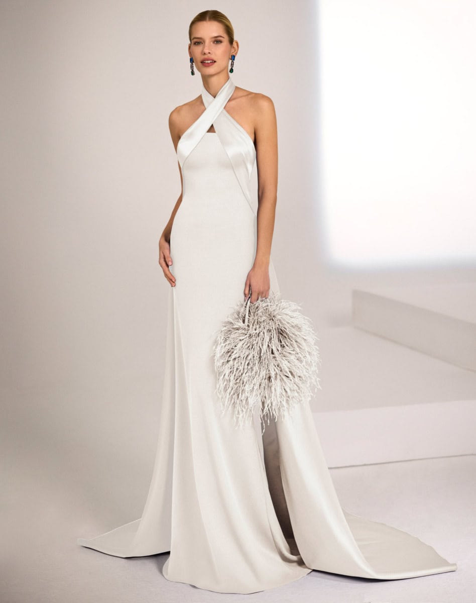 Robe de soirée blanche longue avec décolleté croisé, portée par une femme élégante, accessoirisée avec une pochette en plumes, dans un décor minimaliste.