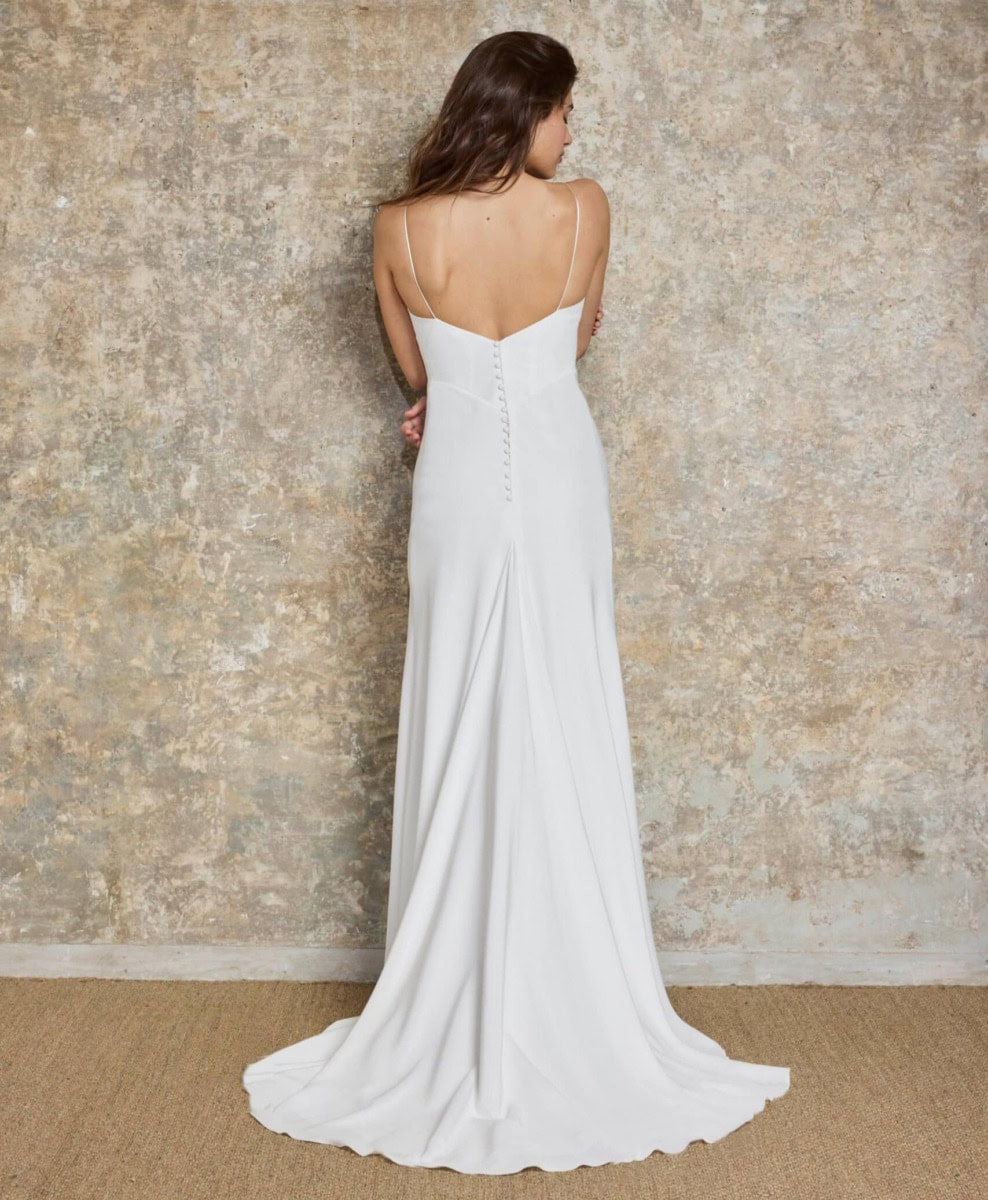 Une femme portant une robe de mariée blanche longue avec dos nu, debout contre un mur en béton brut, vue de dos.