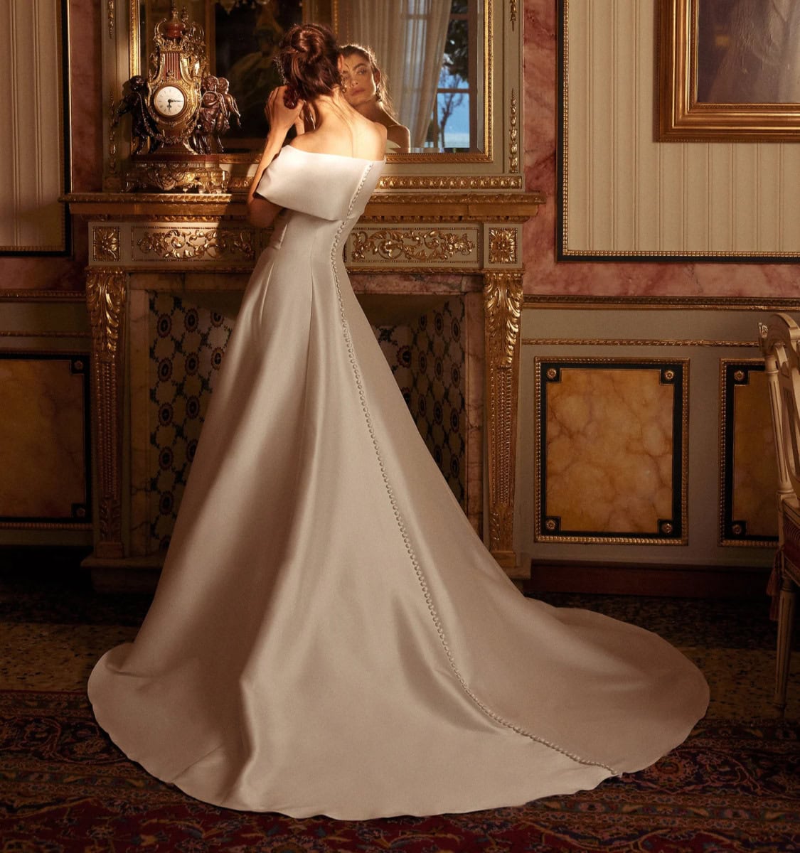Robe de mariée blanche longue avec décolleté épaule, dans un décor vintage riche en détails dorés et meubles anciens, mettant en valeur la beauté et l'élégance de la mariée.