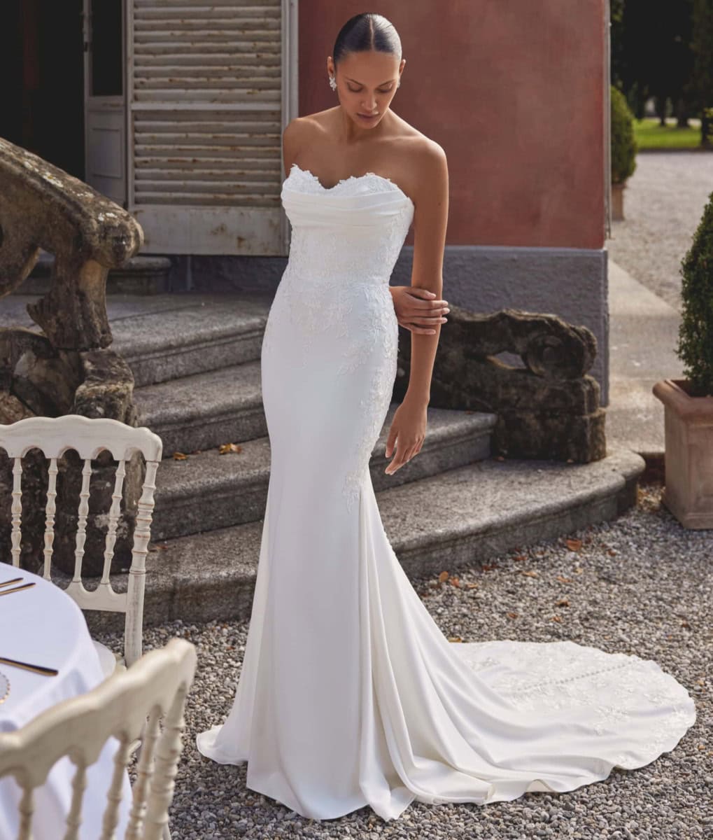 Modèle portant une robe de mariée blanche élégante, ajustée, avec un décolleté en cœur et une traîne longue, dans un cadre extérieur charmant.