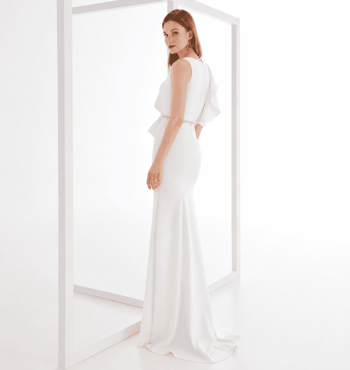 Femme portant une robe blanche élégante avec un design fluide et sophistiqué, vue de côté dans un décor minimaliste blanc.