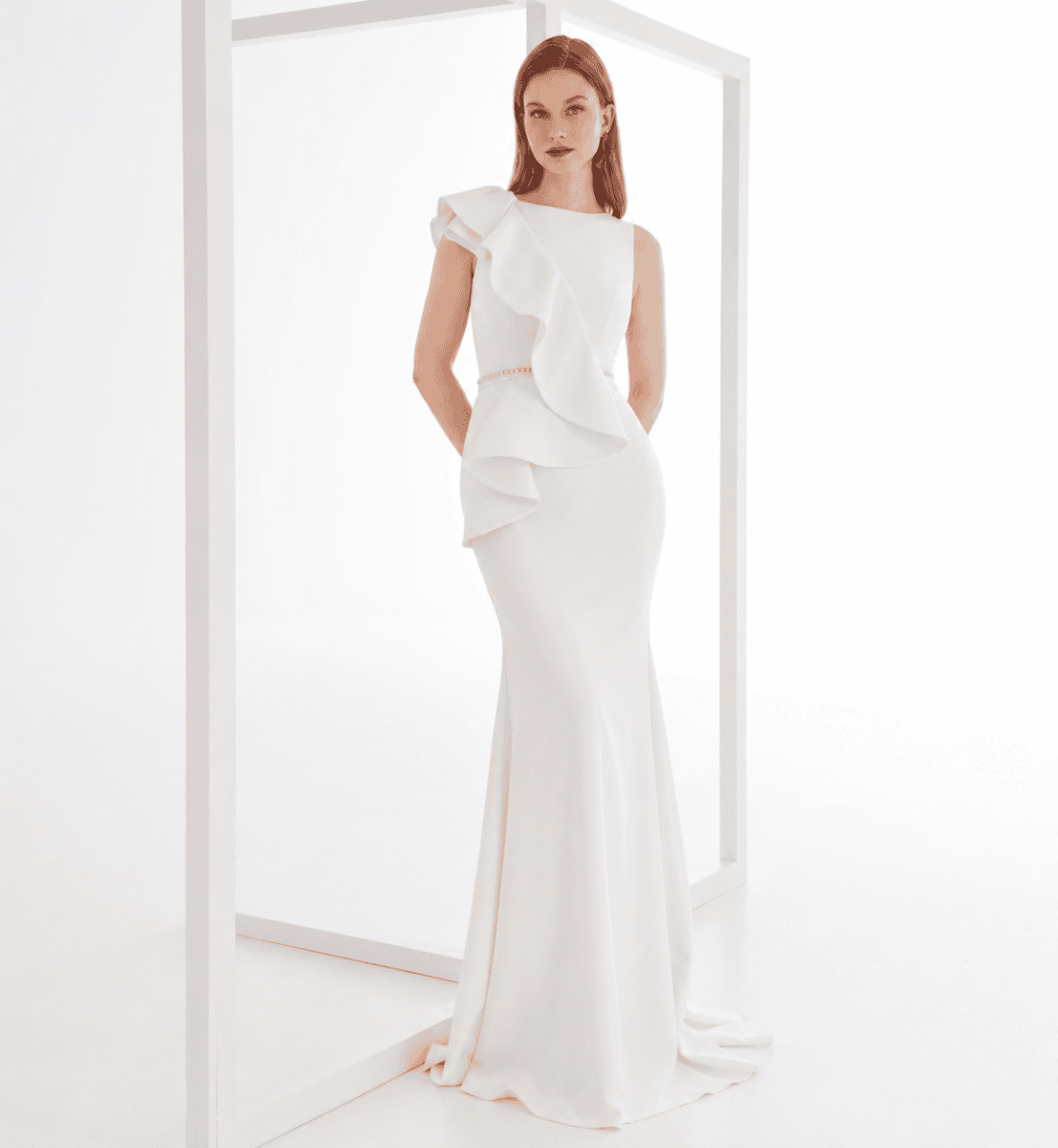Robe de mariée élégante en satin blanc avec détails en volants et taille cintrée, portée par une femme dans un décor minimaliste blanc.