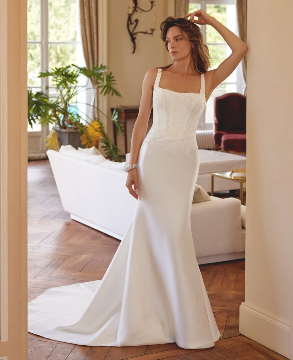 Femme portant une robe de mariée en satin blanc, élégante et sophistiquée, dans un intérieur lumineux avec mobilier classique et plantes vertes.