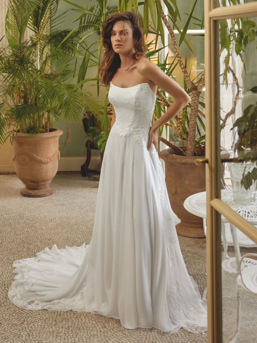 Jeune femme portant une robe de mariée blanche longue et élégante, dans un environnement verdoyant avec des plantes d'intérieur, mettant en valeur le style sophistiqué et romantique.