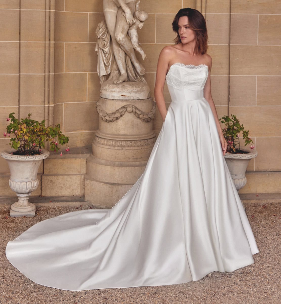 Robe de mariée blanche avec bustier en dentelle et jupe fluide, portée par une femme devant une façade en pierre avec sculptures classiques.