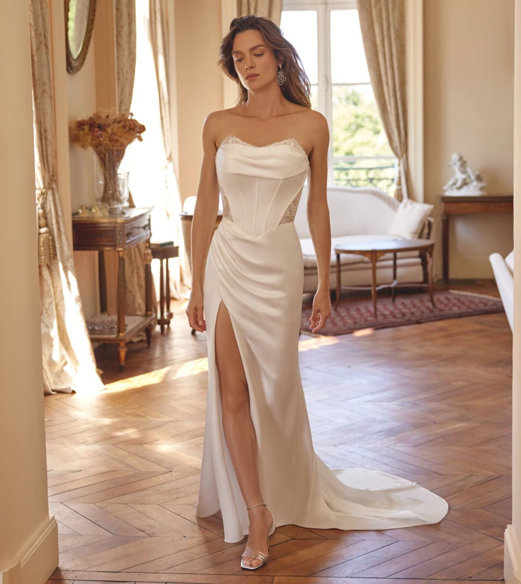 Robe de mariée blanche longue avec décolleté et fente haute, portée par une femme dans une pièce lumineuse, mettant en valeur un style sophistiqué et féminin.
