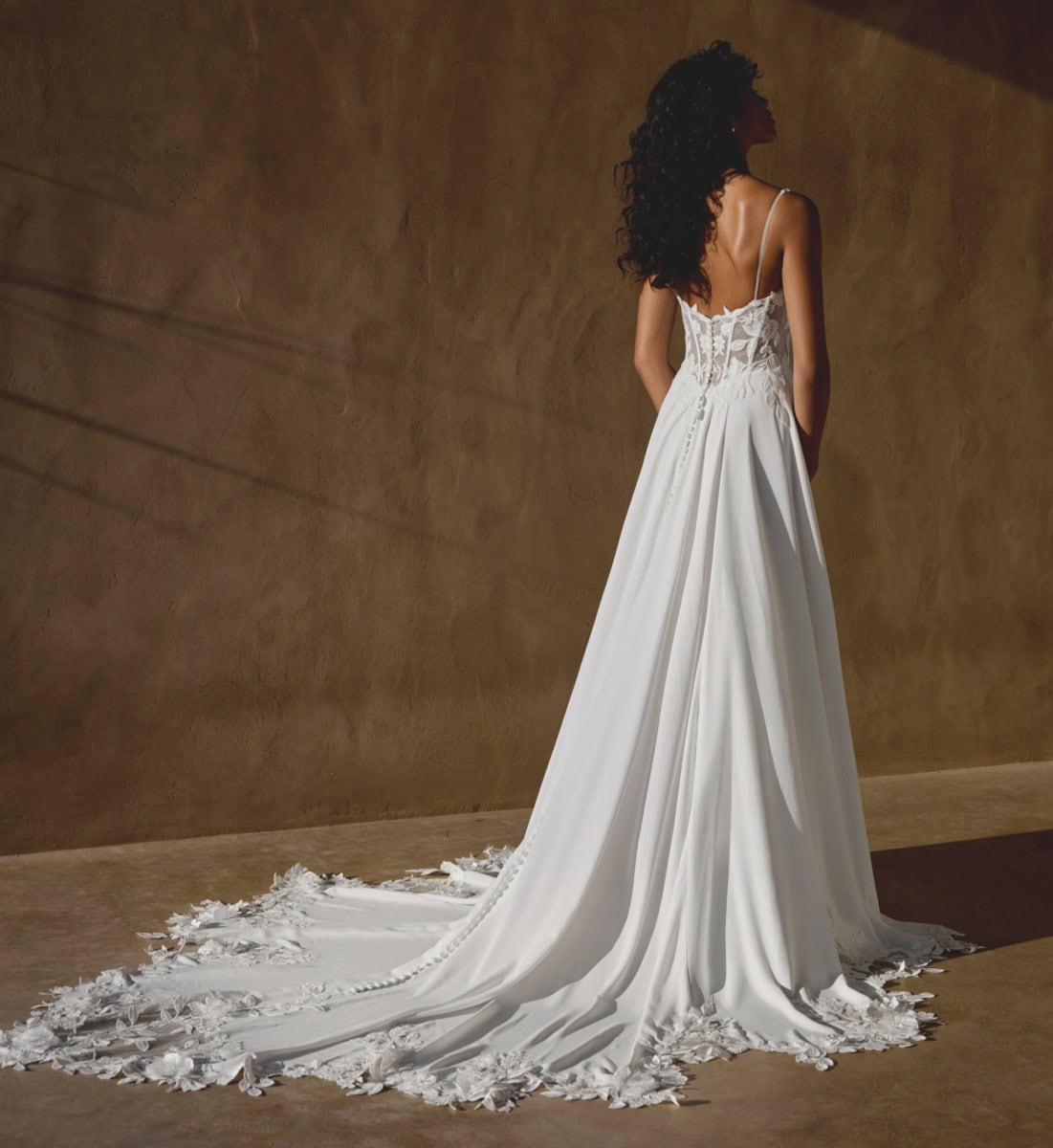Tage de mariage élégants avec une robe longue blanche en satin et dentelle, portée par une femme aux cheveux bouclés, dans un décor minimaliste avec un mur beige.