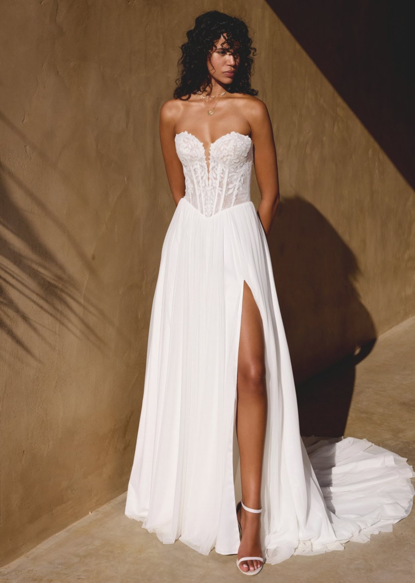 Une femme portant une robe de mariée blanche élégante avec un corsage en dentelle et une jupe fluide à fente, posant devant un mur beige avec un éclairage doux.