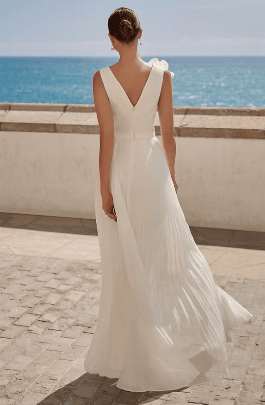 Femme en robe blanche longue avec épaules dénudées regardant la mer depuis une promenade en bord de mer.