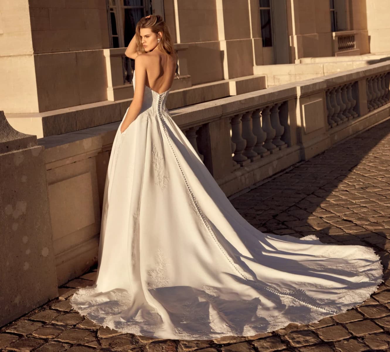 Robe de mariée élégante avec corset en dentelle et longue traîne, portée par une femme dans un décor urbain en fin d'après-midi.
