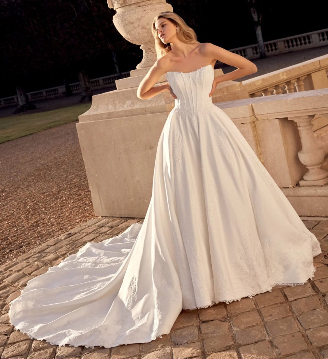 Robe de mariée blanche élégante avec corsage plissé et longue traîne, portée par une femme dans un décor extérieur historique.