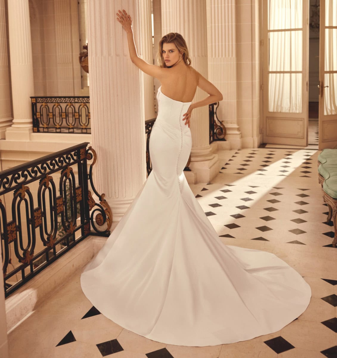 Femme en robe de mariée blanche élégante dans un intérieur classique avec colonnes et balustrades en fer forgé.