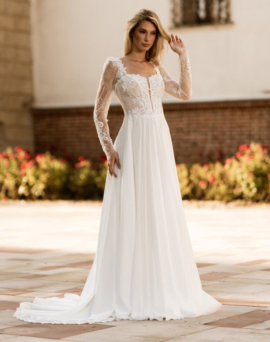 Léa portant une robe de mariée élégante avec dentelle, manches longues et jupe fluide, posant en extérieur dans un cadre ensoleillé avec des fleurs colorées en arrière-plan.