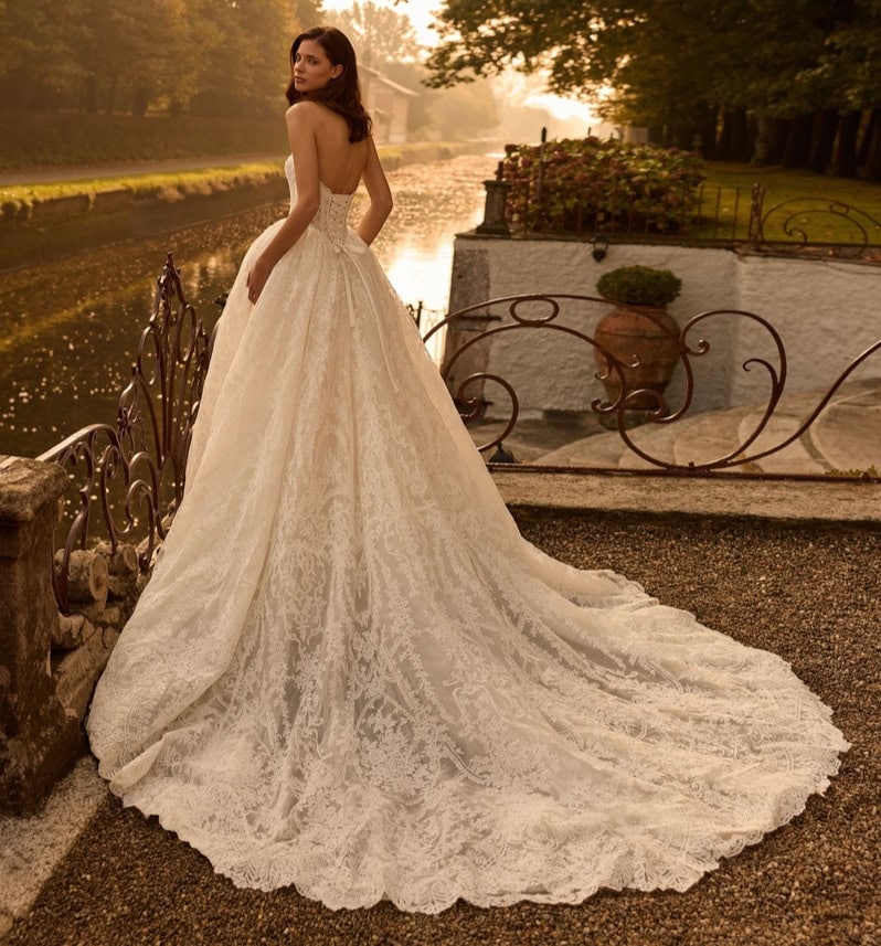 Elegant wedding dress with lace details, flowing train, and strapless design, set outdoors near a river at sunset, perfect for bridal fashion and special occasions.