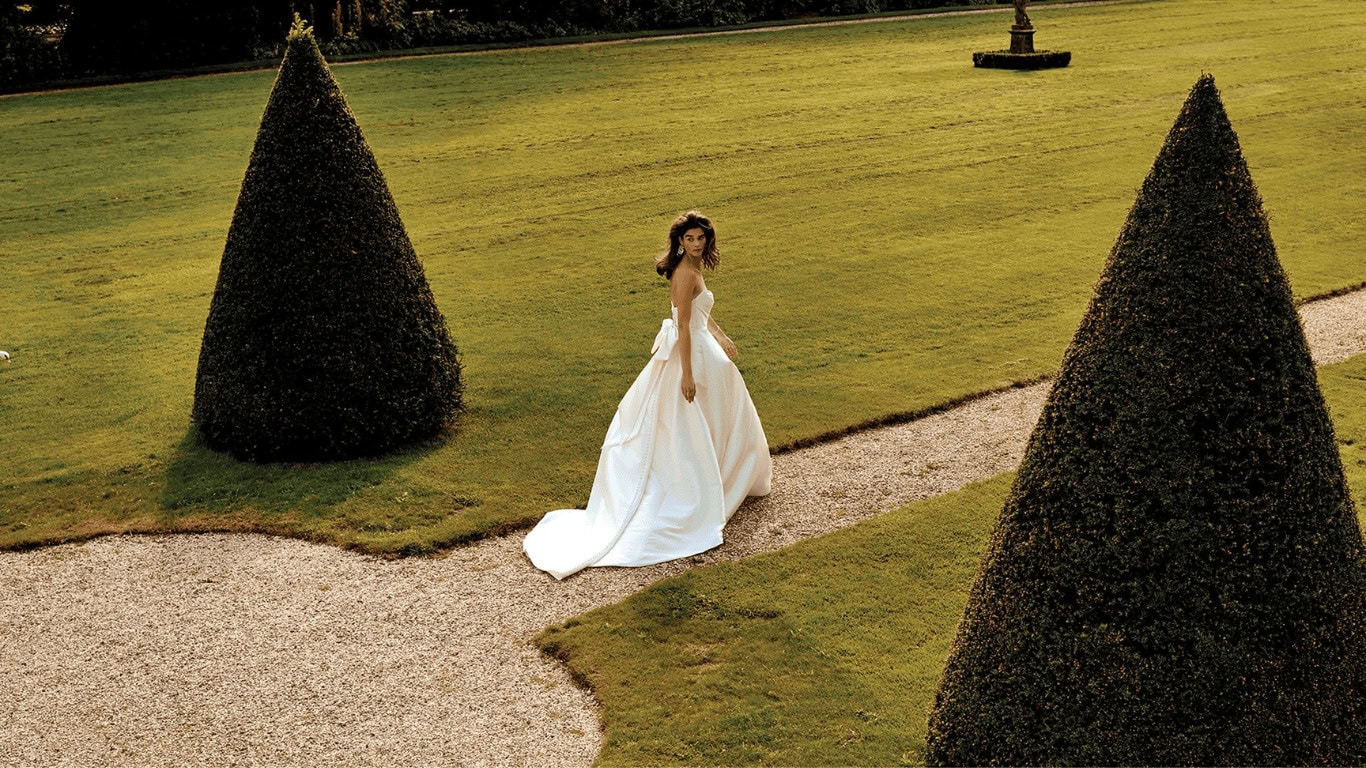 Femme en robe de mariée blanche dans un jardin bien entretenu avec des topiaires coniques et un chemin de gravier.