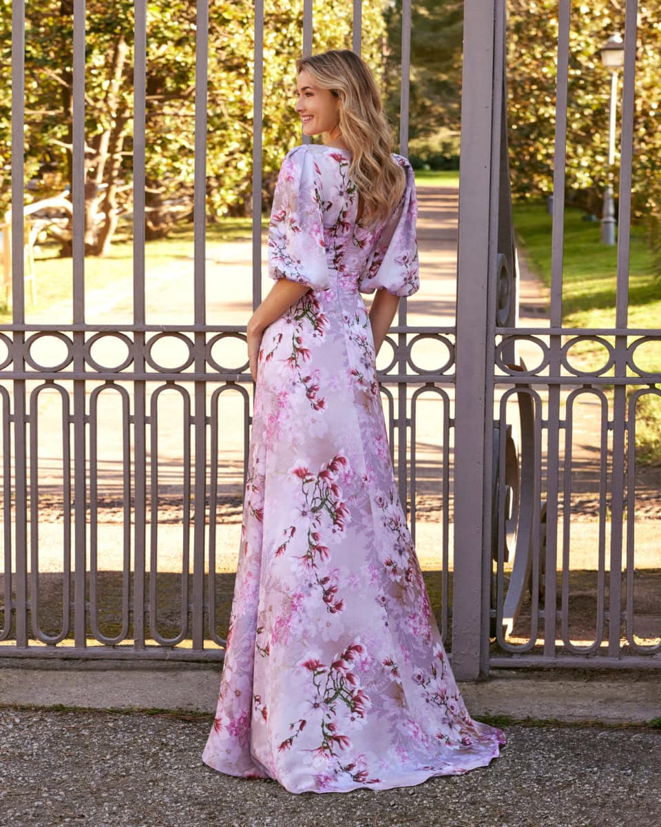 Femme portant une robe longue en satin rose avec motifs floraux, debout devant une grille en fer forgé dans un parc ensoleillé.
