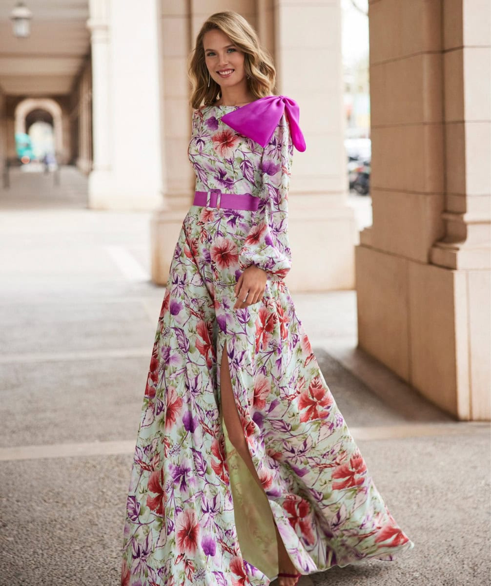 Femme portant une robe longue florale avec un grand nœud violet sur l'épaule dans un environnement urbain élégant.
