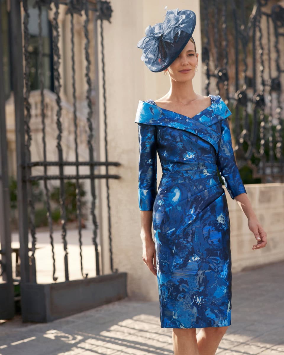 Femme élégante portant une robe bleue avec un chapeau assorti, posant devant une grille en fer forgé dans un cadre urbain.