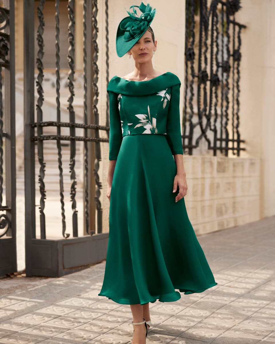 Femme élégante portant une robe verte longue avec un chapeau assorti, posant devant une grille en fer forgé à l'extérieur.