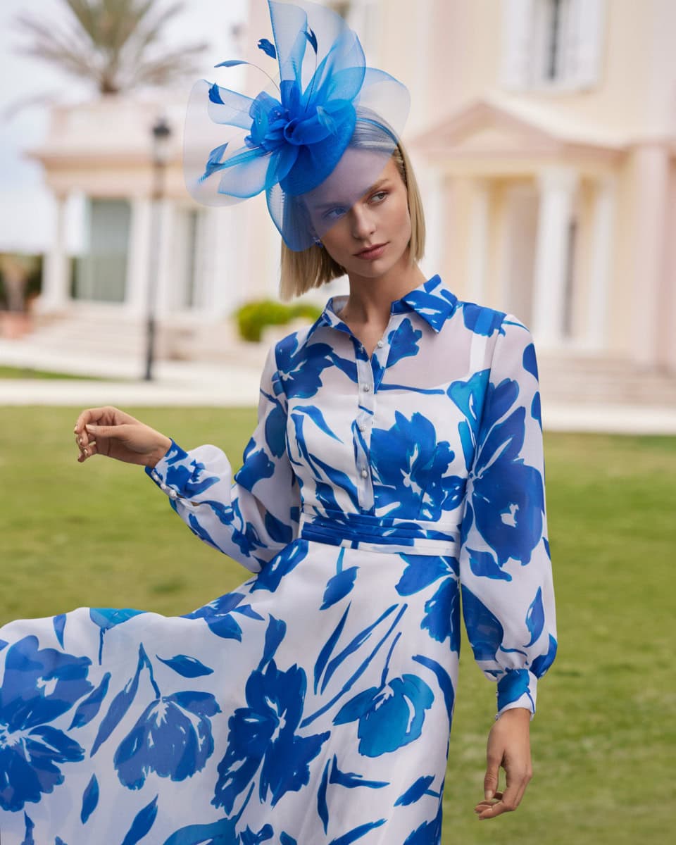 Modèle portant une robe blanche à motifs bleus avec un grand chapeau en tulle bleu, devant une maison élégante.