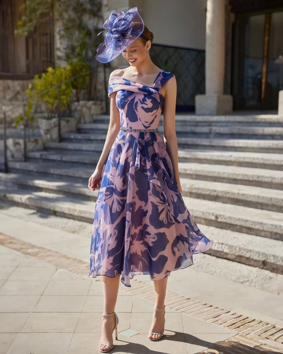 Femme élégante portant une robe longue à motifs floraux bleus et roses, avec un chapeau assorti, en extérieur devant un bâtiment historique.