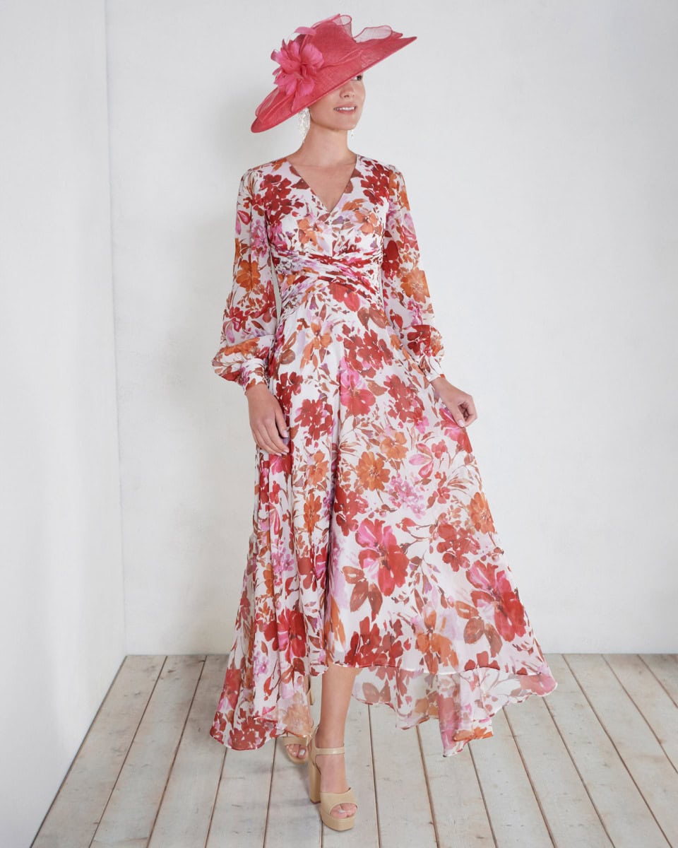 Femme élégante portant une robe longue à fleurs roses et orange avec un grand chapeau rose, debout contre un mur blanc.