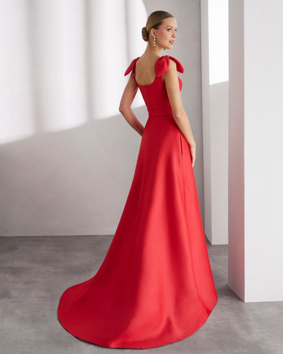 Robe de soirée rouge élégante avec épaules à volants, longue traîne, portée par une femme dans un intérieur moderne.