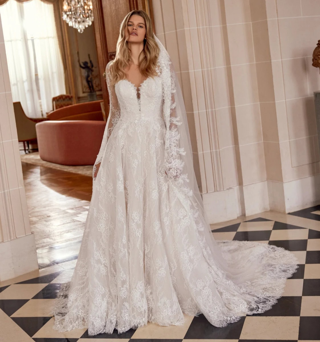 Robe de mariée élégante en dentelle blanche avec manches longues et décolleté en V, portée par une femme dans un intérieur sophistiqué.